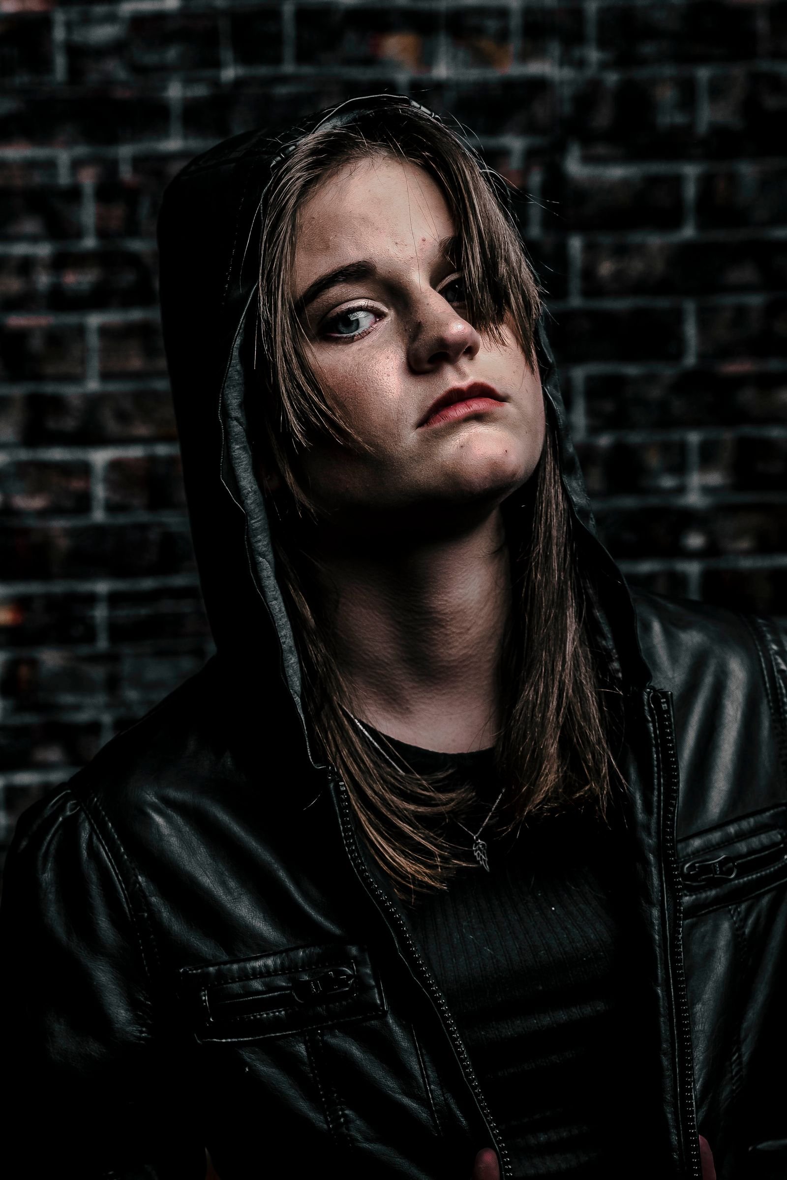 Moody hooded portrait in leather against a brick wall, low light